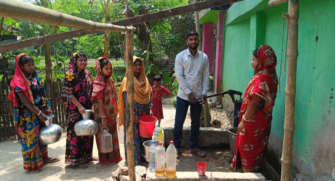 Women waiting at a village well with bottles of pale yellow water on the ledge